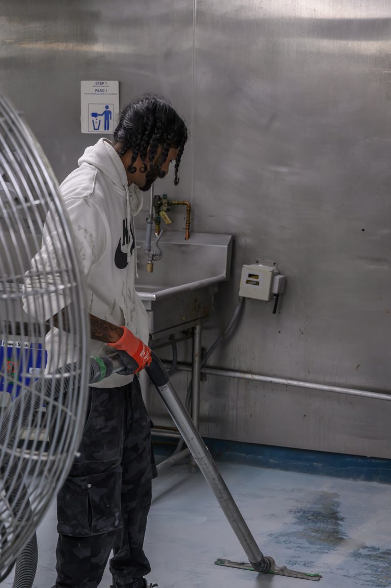 A person in a white hoodie uses a vacuum attachment to clean a stainless steel kitchen floor.