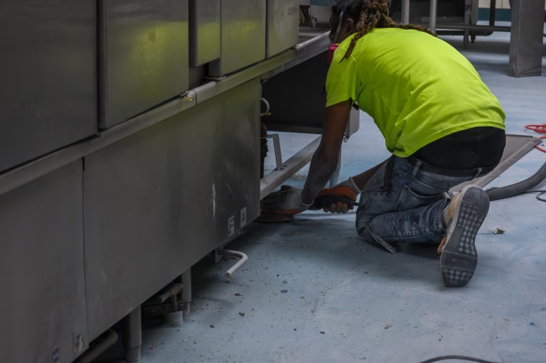A worker in a lime-green shirt kneels to sand the base of a large stainless steel industrial machine.