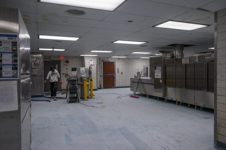 A worker cleans the floor of a commercial kitchen near a large stainless steel dishwasher.
