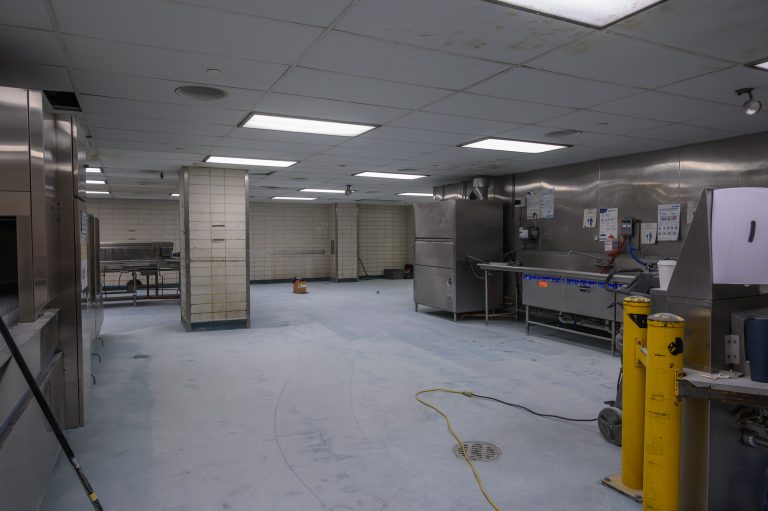 Empty commercial kitchen featuring stainless steel equipment and a pale, dusty floor.