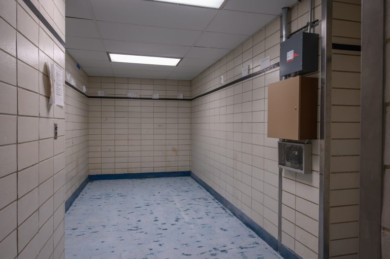 An empty hallway with beige tiled walls, a blue floor, and wall-mounted electrical utility boxes.