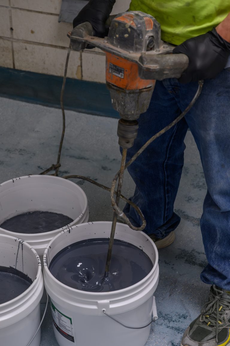 A worker in black gloves uses a power drill and paddle to stir gray paint in a bucket.