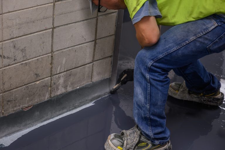 A worker in a high-visibility vest brushes gray epoxy coating onto a floor edge.