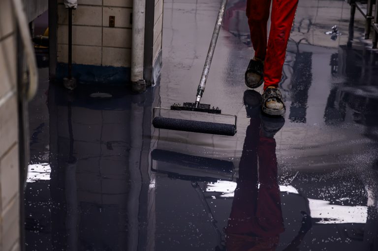 A worker in red pants rolls dark gray epoxy coating onto a floor.