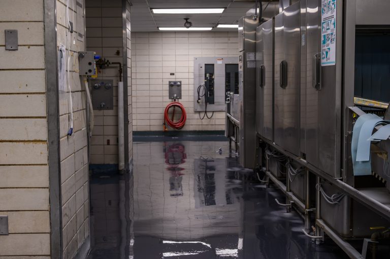 A commercial kitchen features a large stainless steel dish machine beside a red fire hose on tiled walls.