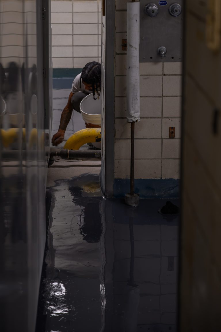 A person kneels on a wet floor in a tiled shower, holding a bucket while working.