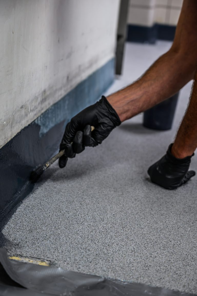 A person in black gloves uses a brush to apply gray coating along the edge of a floor.