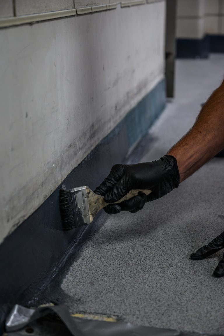 A person in black gloves uses a brush to paint a gray border along the floor and wall.