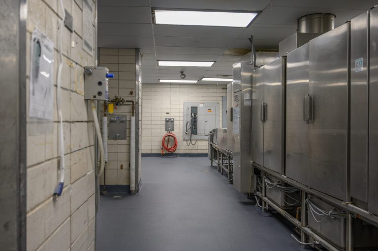 Stainless steel industrial kitchen appliances line a tiled room with a red wall-mounted hose.
