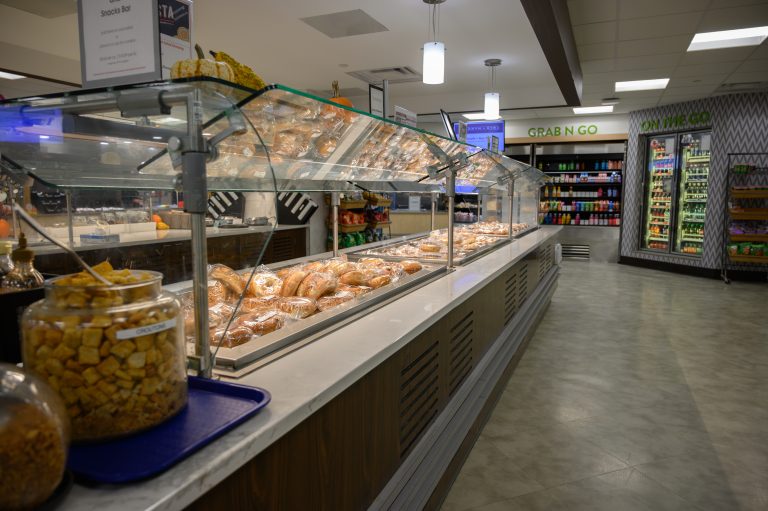 A self-serve food station features wrapped bread rolls, a jar of croutons, and grab-and-go coolers.