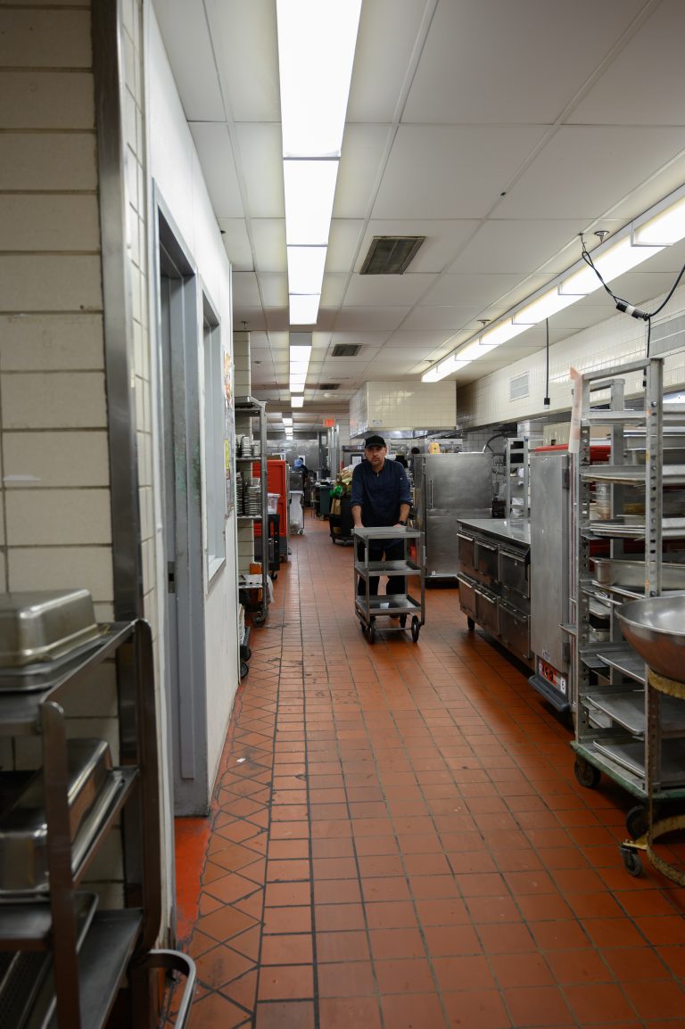 A chef in a uniform pushes a metal cart through a professional commercial kitchen.