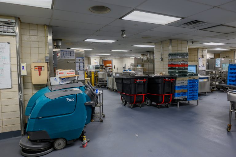 A teal floor scrubber is parked in a commercial kitchen beside rolling supply bins and plastic crates.