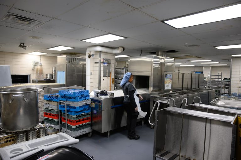A kitchen worker loads trays into a large industrial dishwasher in a commercial kitchen.